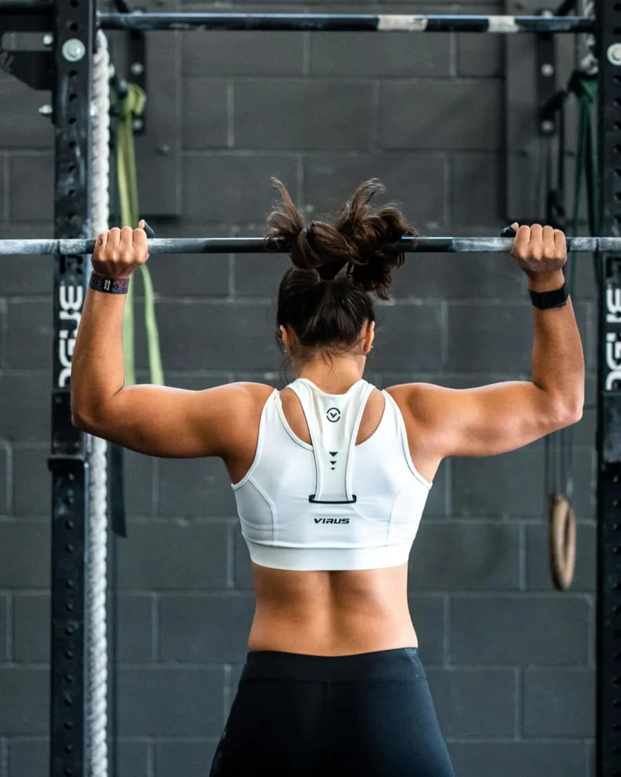 A figure pressing a loaded barbell overhead in a dim training hall, seen from behind.