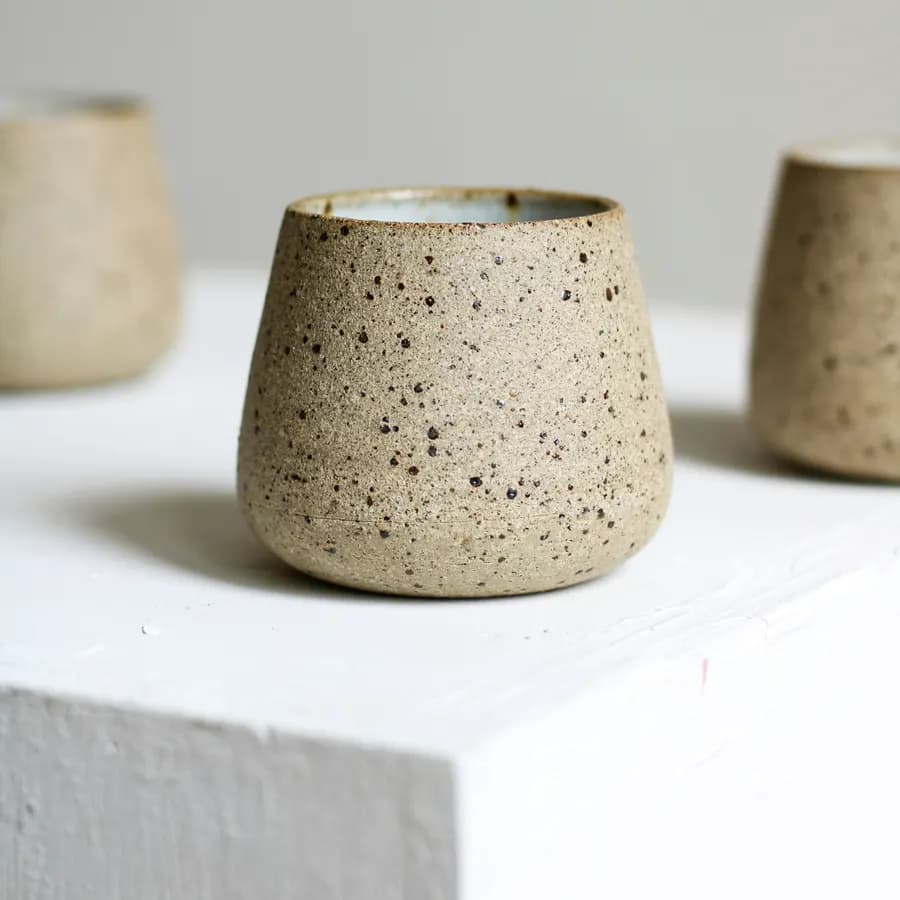 A speckled stoneware cup with a raw clay body, lit from the side on a pale plinth.