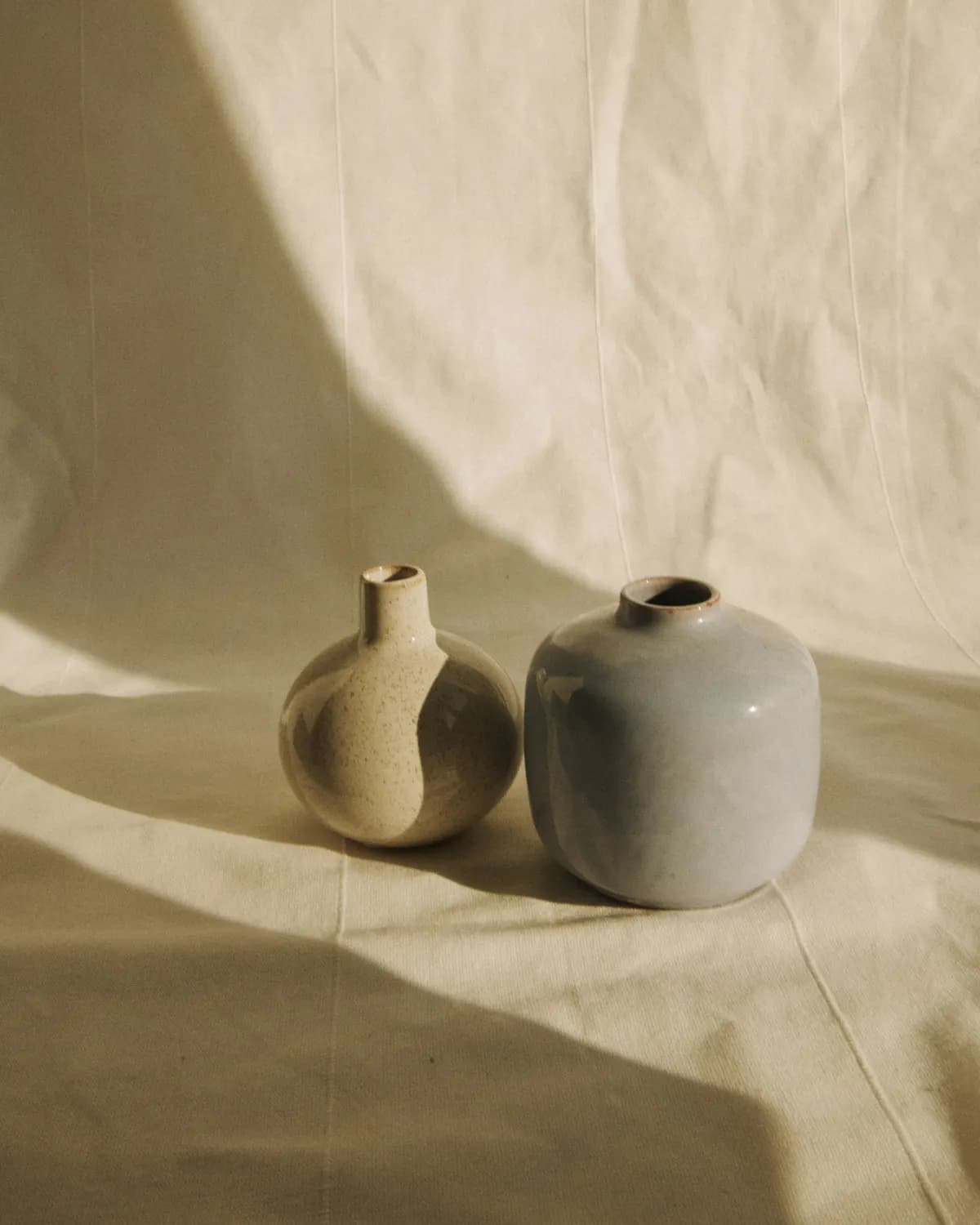 A speckled and a sage-glazed vase resting on folded linen, raked by low afternoon light.