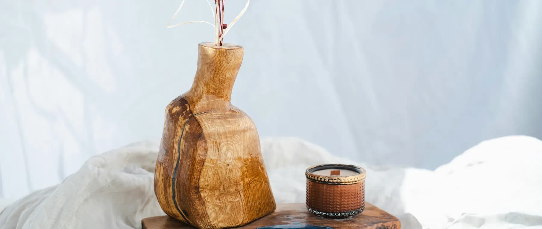 A carved olive-wood vase and a small candle on a live-edge board, lit against white linen.