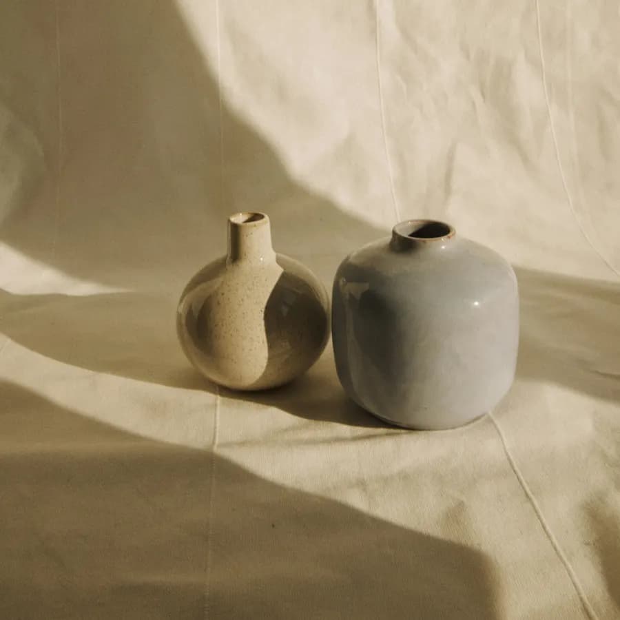 A speckled and a sage-glazed vase side by side, shadows falling across folded linen.