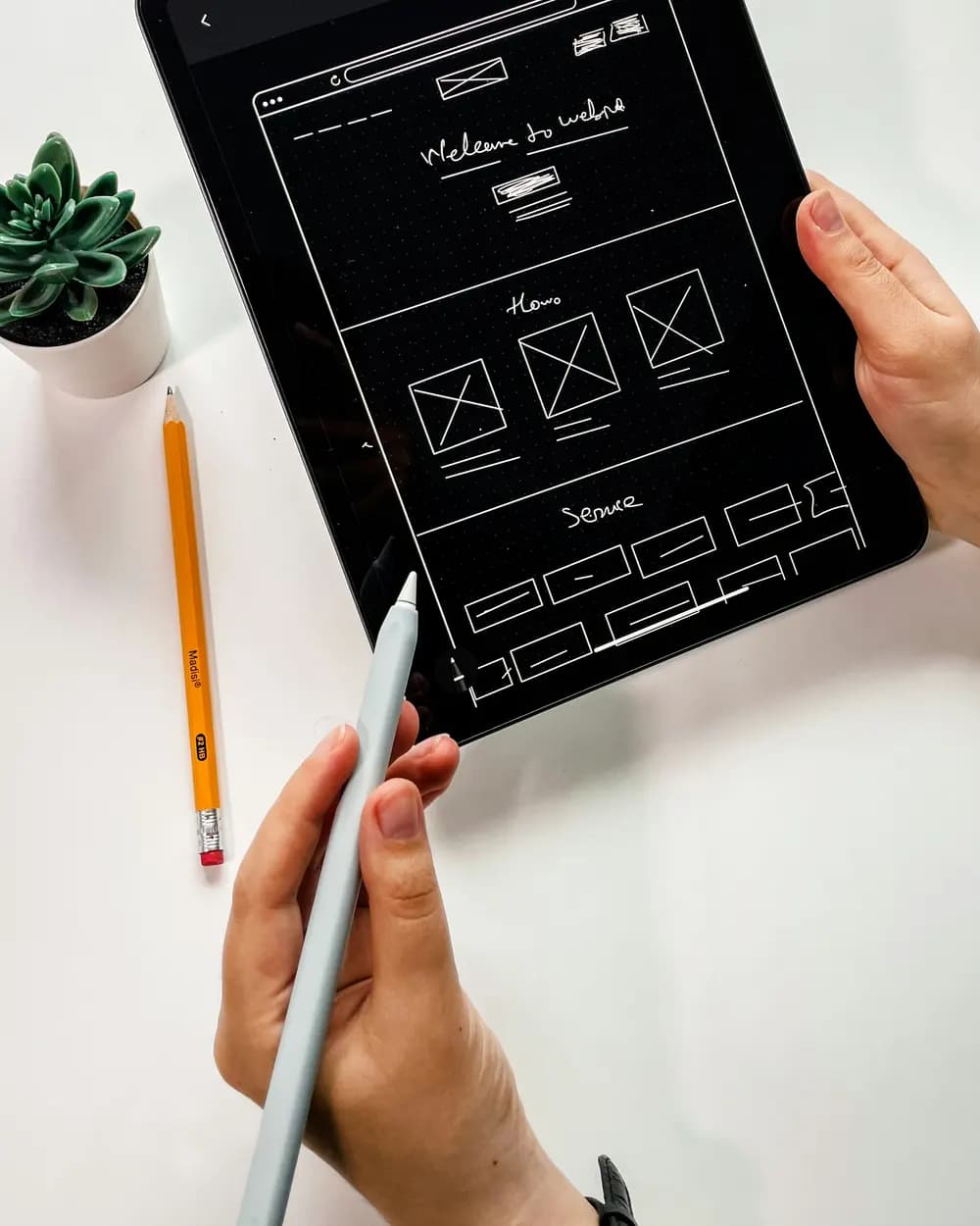 Hands sketching a page wireframe on a tablet with a stylus, beside a pencil and a plant.